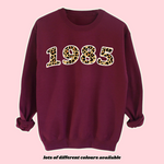 Load image into Gallery viewer, Retro birth year sweatshirt with leopard print numbers on a burgundy fleece-lined jumper. Vintage 80s and 90s style personalised birthday sweatshirt, perfect for milestone birthdays like 30th, 40th, or 50th. Unisex oversized fit, customisable colours and text. Trendy throwback fashion gift idea
