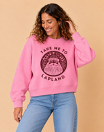 Load image into Gallery viewer, A woman wearing a pink crewneck sweatshirt with a dark brown graphic of a bearded man and the text 'TAKE ME TO LAPLAND' and 'FINLAND', paired with light blue jeans, set against a solid beige background.
