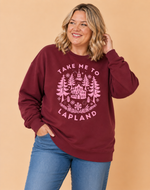 Load image into Gallery viewer, A woman wearing a maroon crewneck sweatshirt with a pink graphic that reads 'TAKE ME TO LAPLAND' and shows blue jeans, set against a solid beige background.
